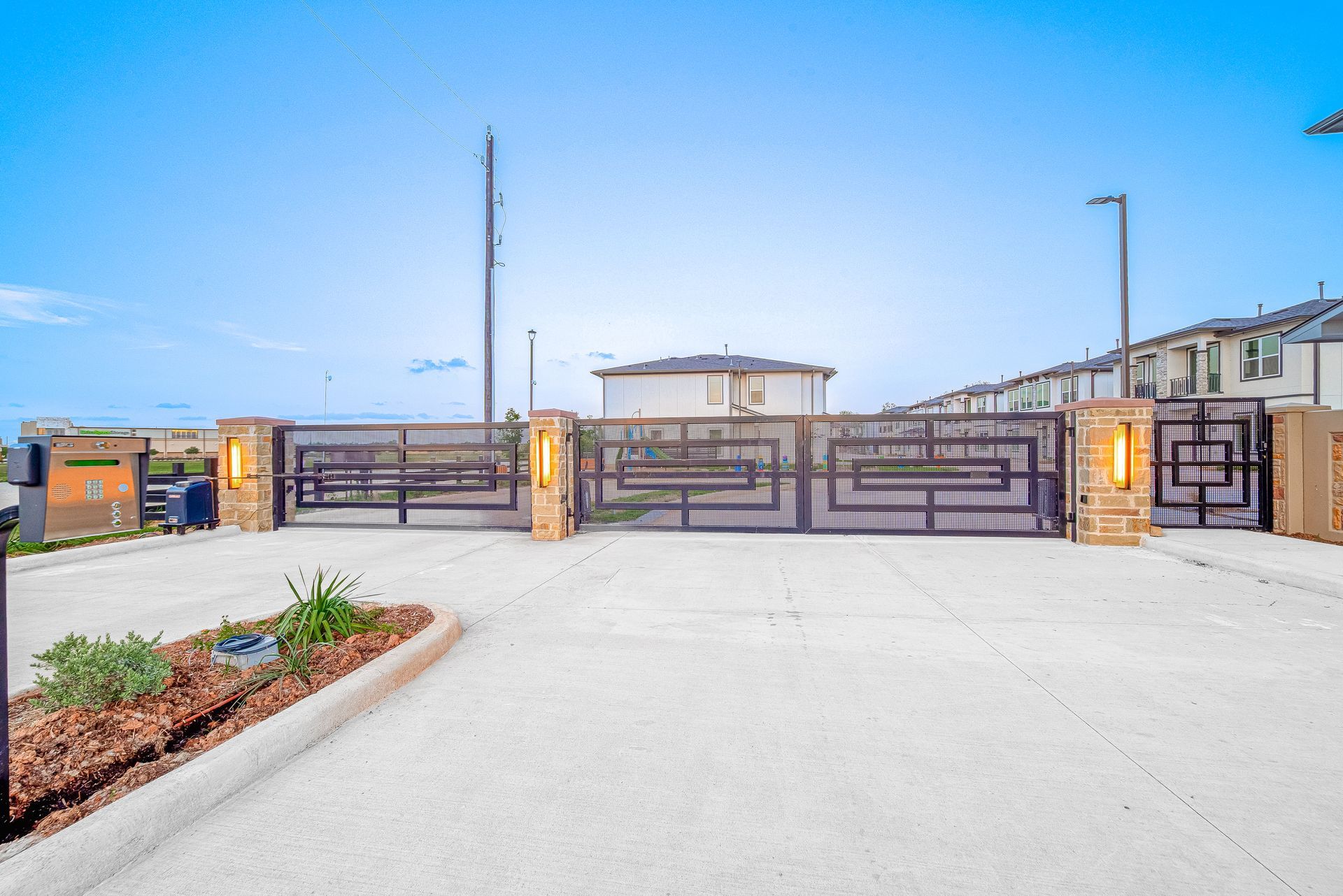Modern gated residential entrance with wide driveway and metal fence under a blue sky