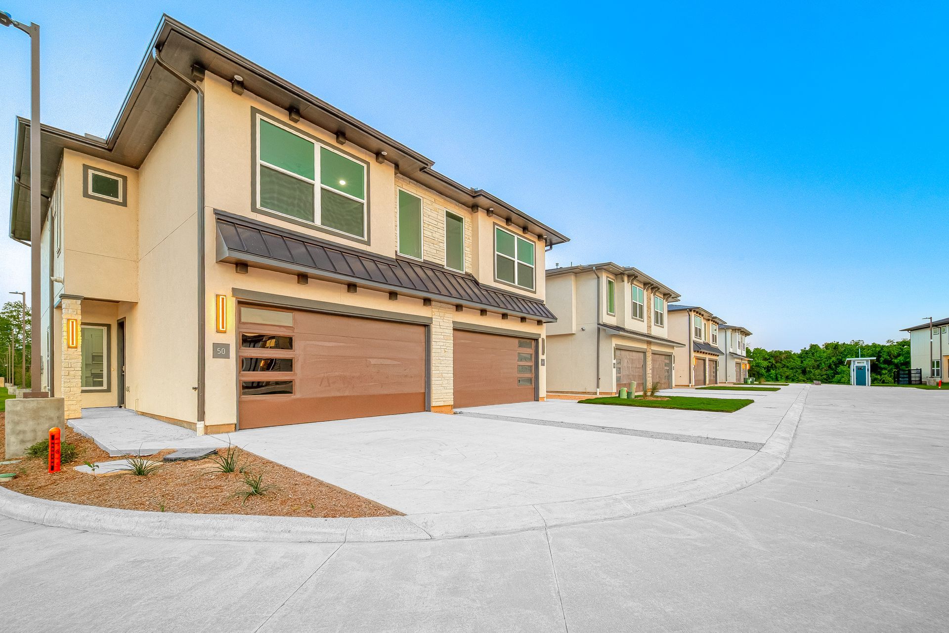 Modern suburban townhouses with tan stucco, garage doors, and a wide empty street under a clear blue sky