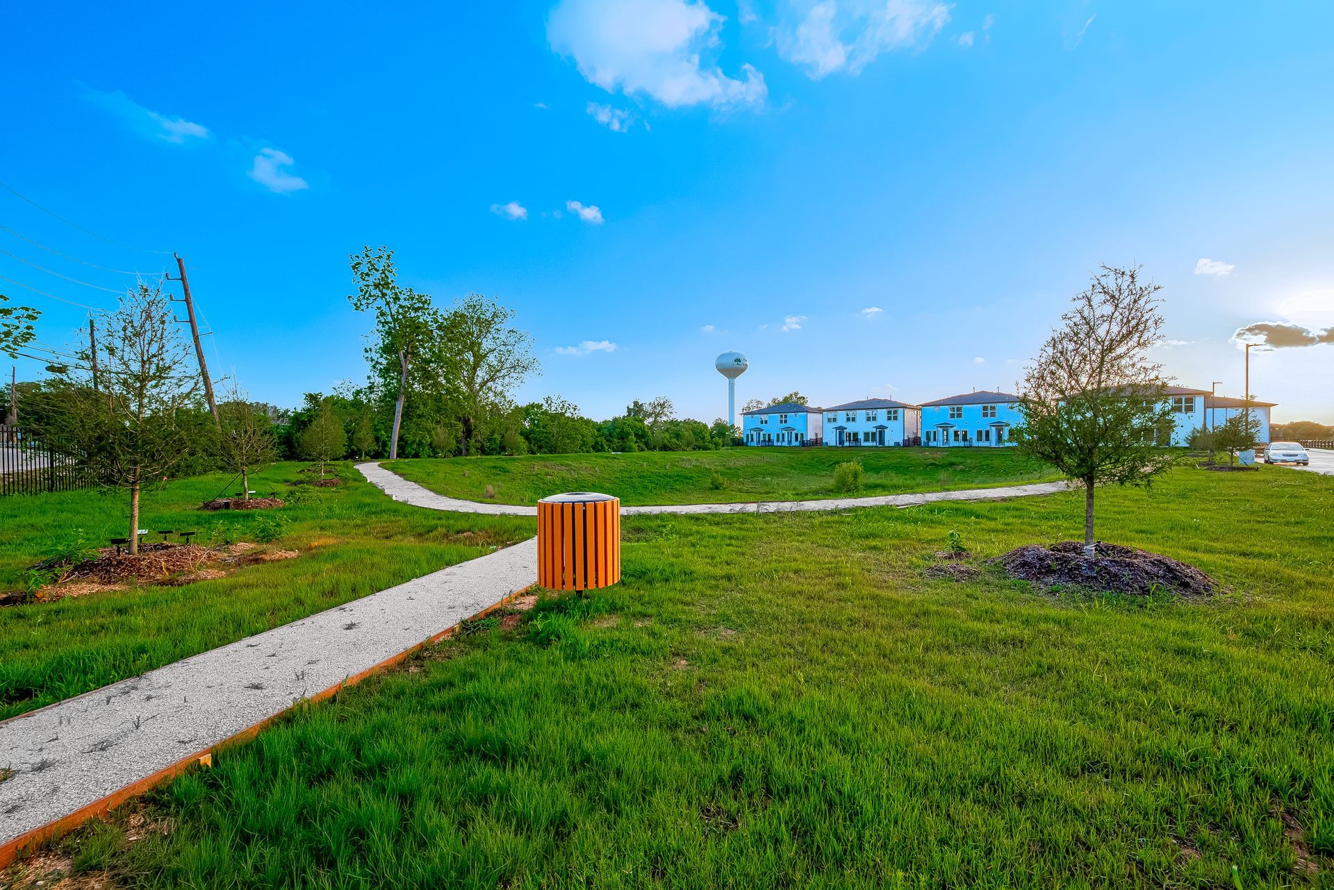 Green park with winding gravel path, grassy lawn, young trees, and blue houses under a bright sky