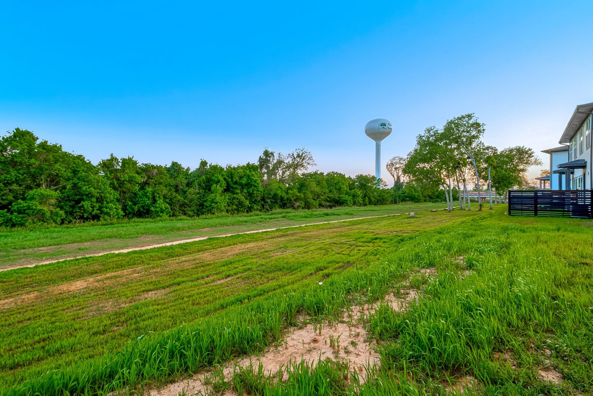 Grassy field with dirt tracks, trees, and a water tower under a bright blue sky