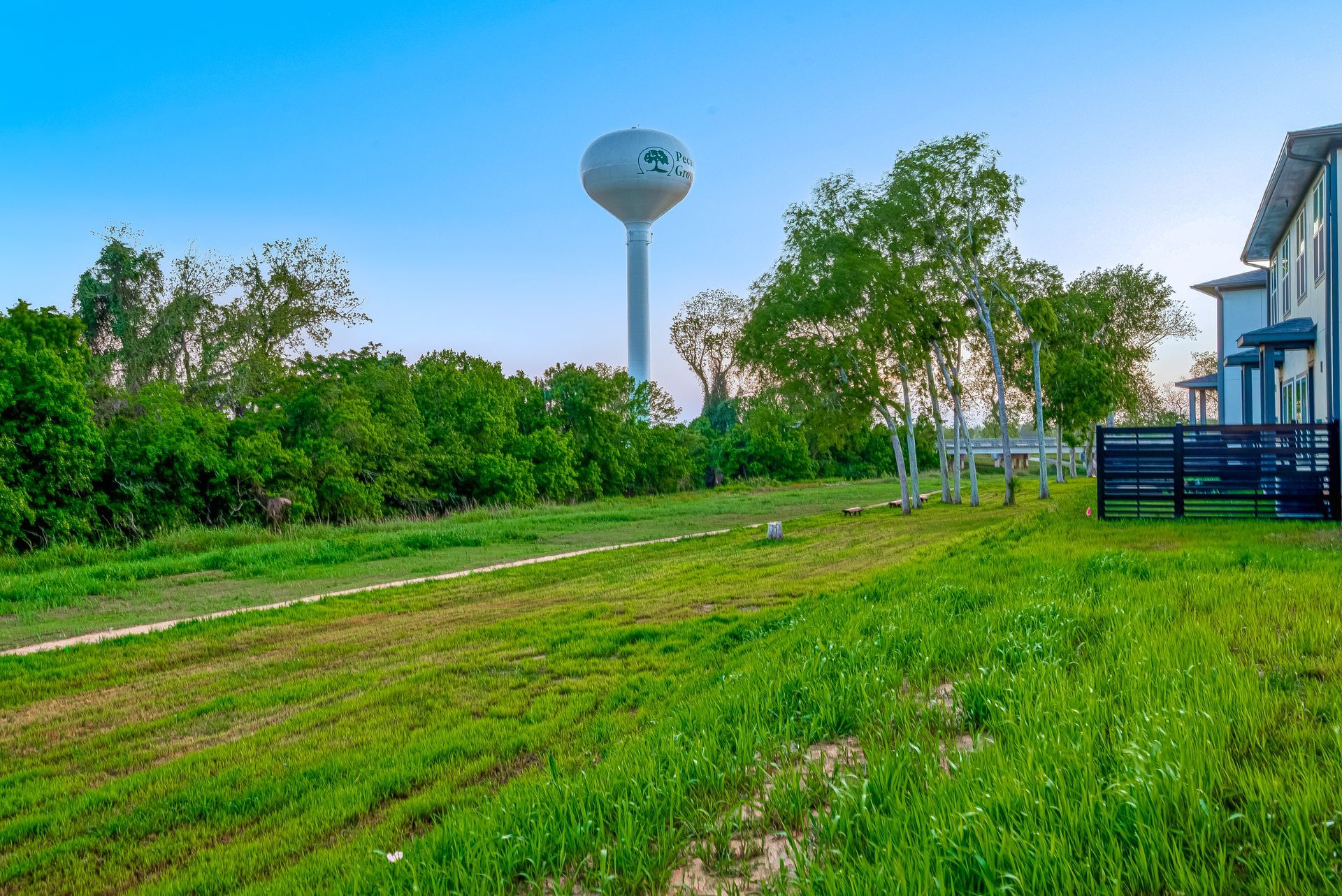 Green grassy field beside a house, with trees and a distant water tower under a blue sky.