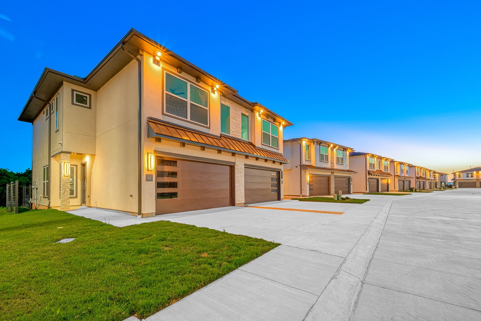 Row of new beige townhomes with garage doors, lit at dusk beside a wide sidewalk and green lawn