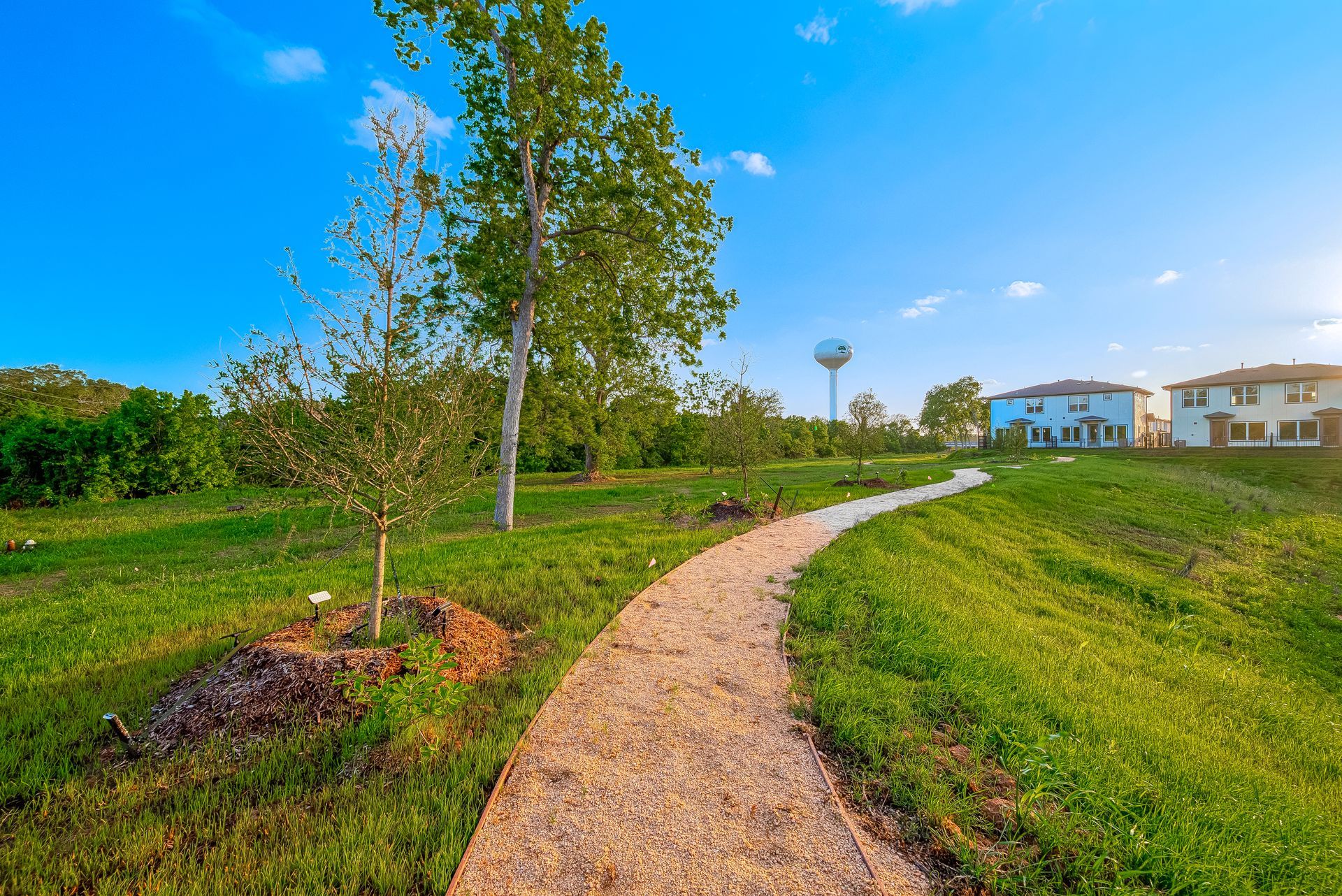 Curving gravel path through a green park toward houses under a blue sky