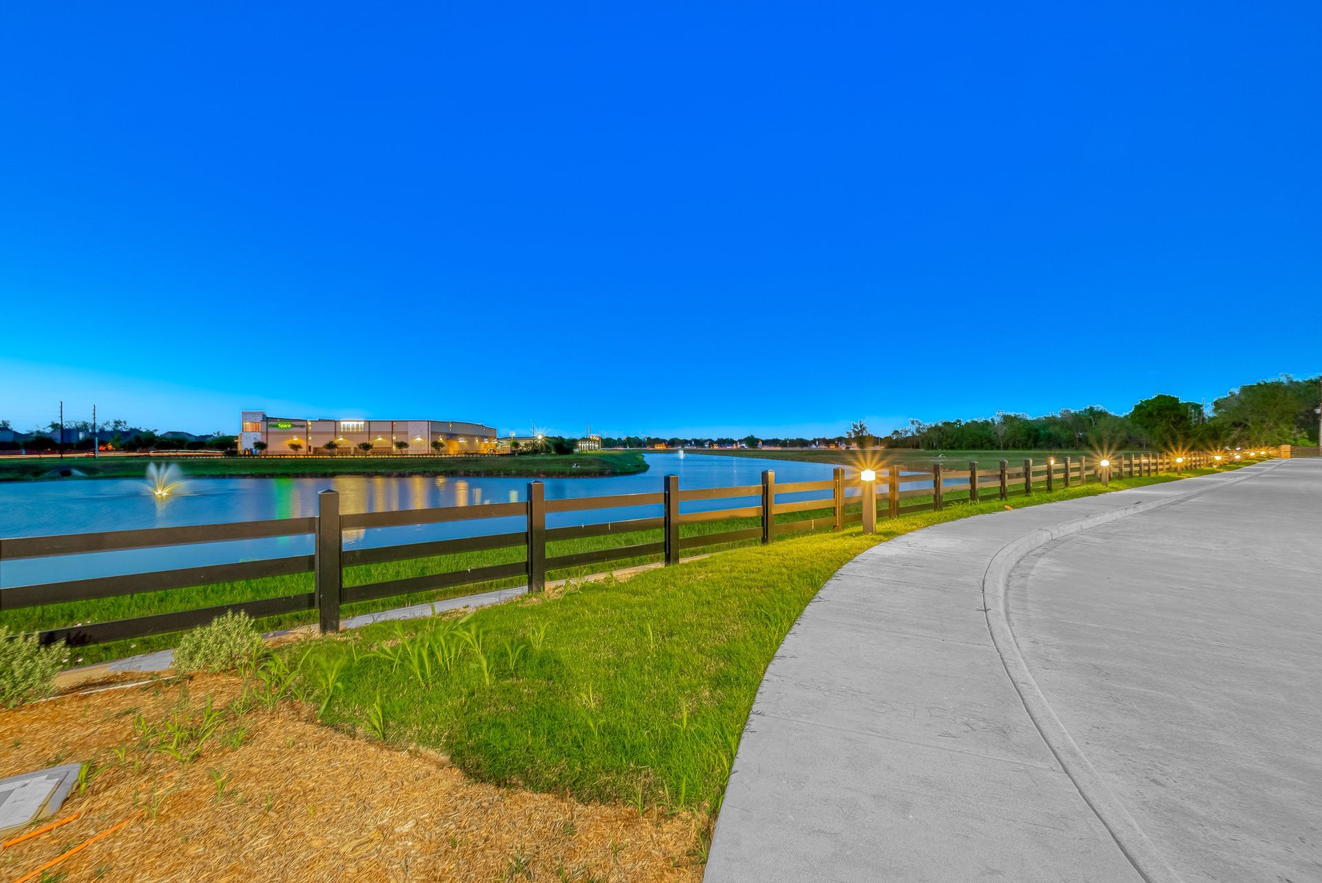 Curved riverside path beside a wooden fence, grassy bank, and illuminated buildings at dusk under a blue sky