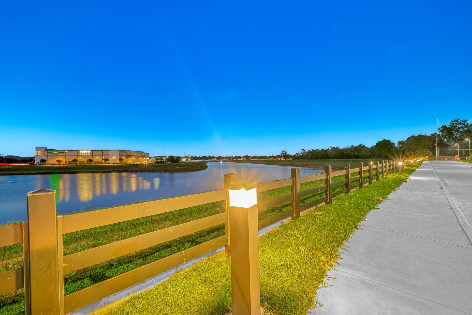 Lit riverside path with wooden fence at dusk under a bright blue sky