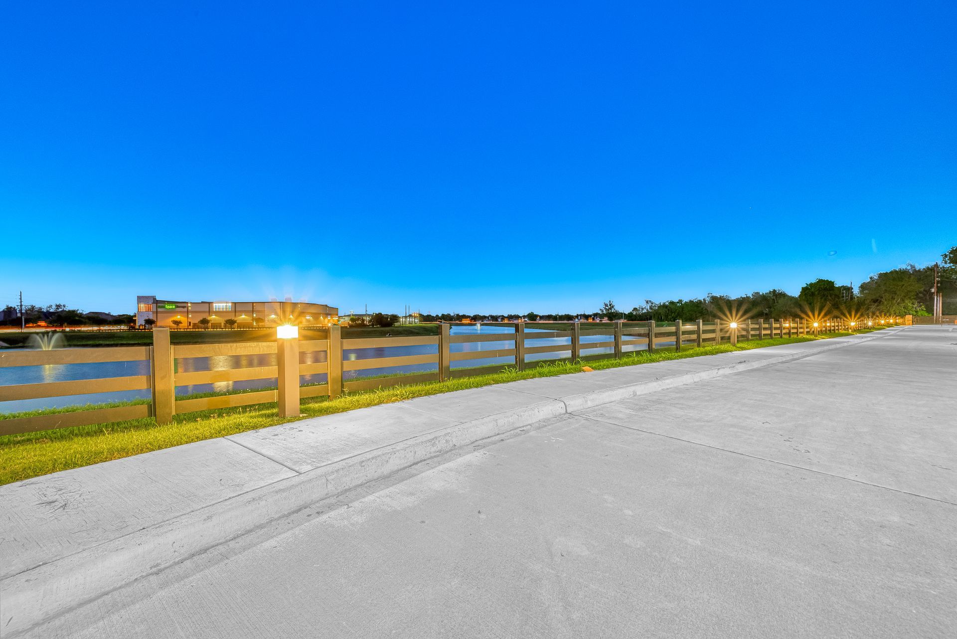Sunset over a waterfront road with a wooden fence and glowing lights along the shore