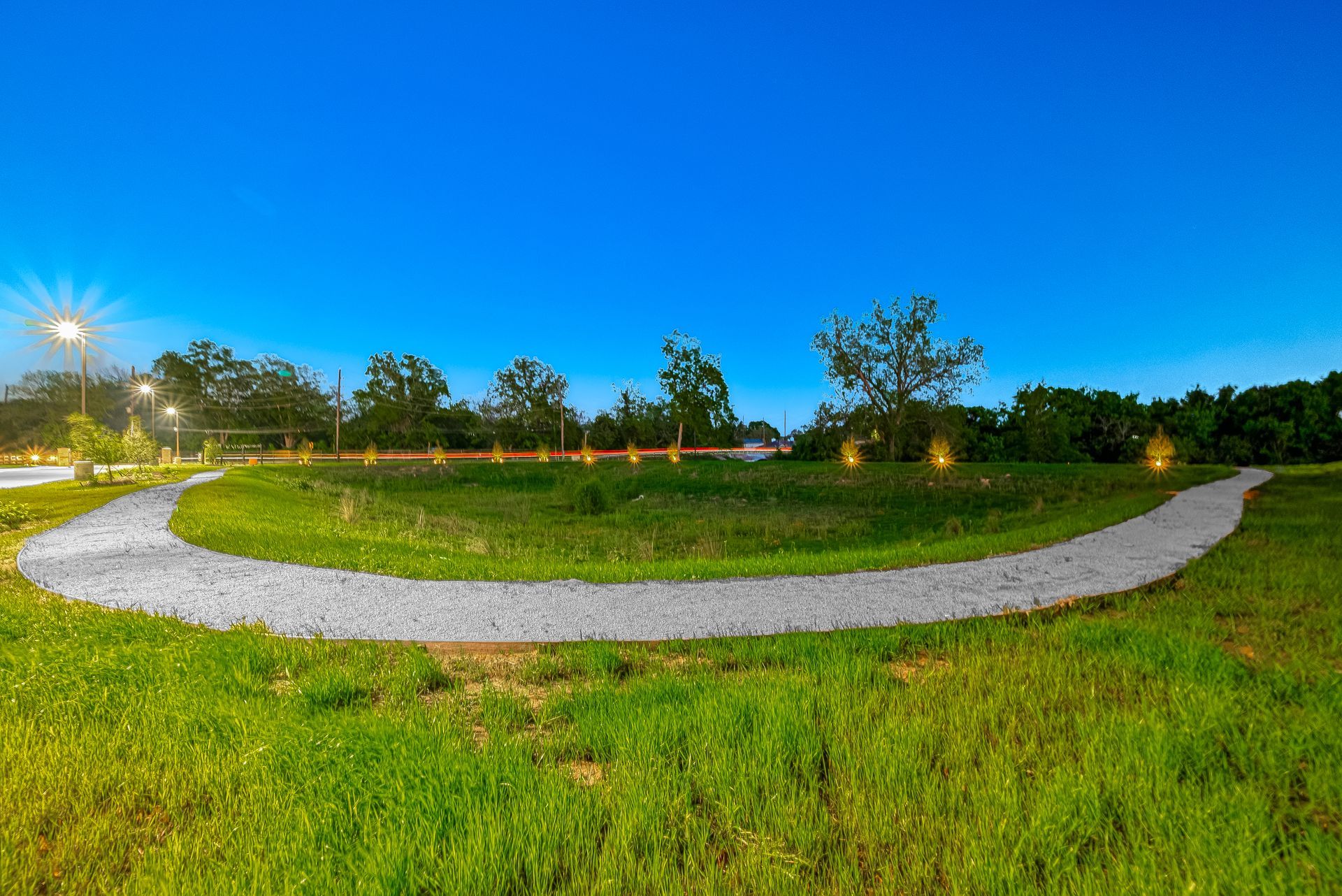 Curved gravel path through a grassy park at dusk under a blue sky with scattered trees and lights