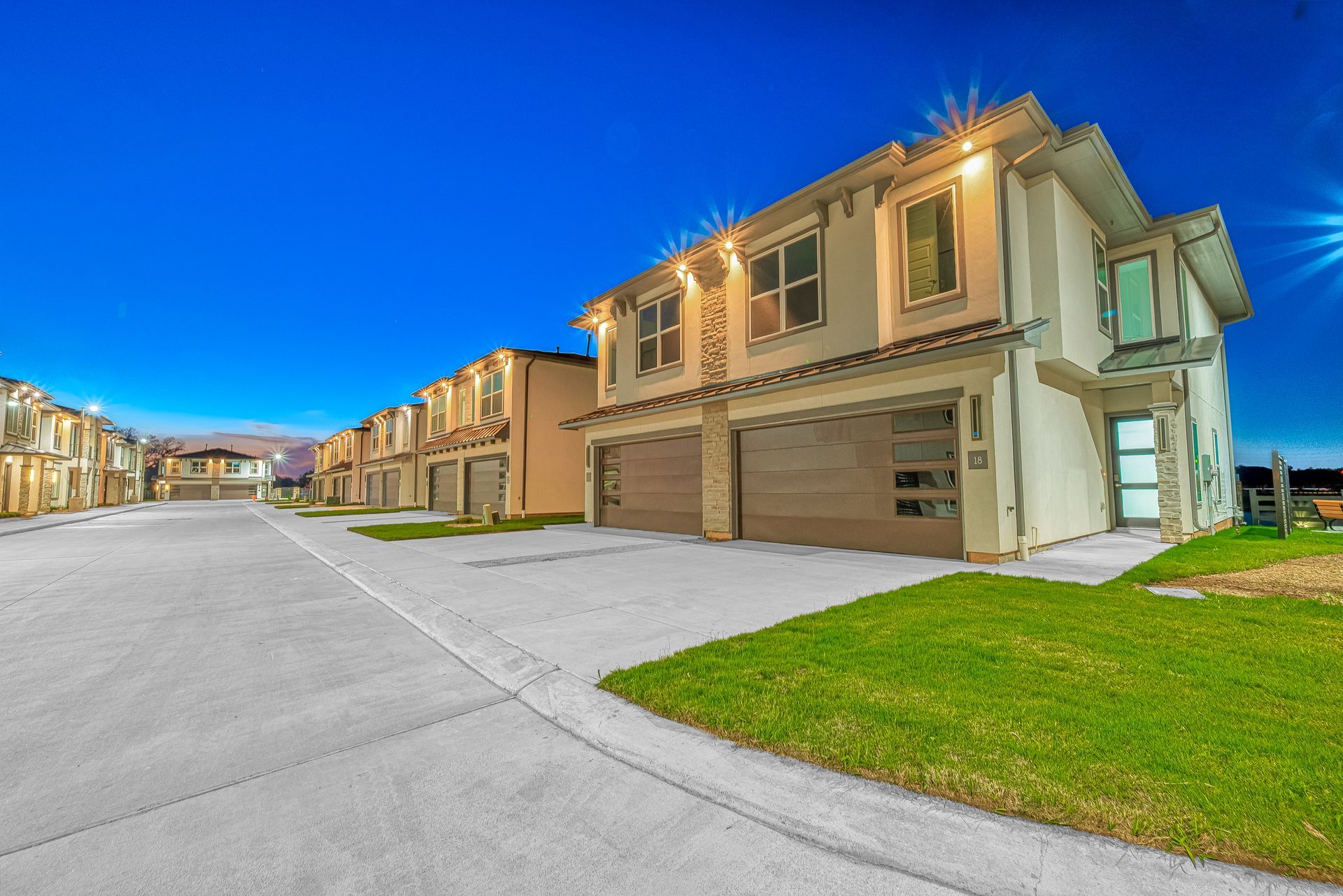 Row of modern suburban townhouses at dusk, with beige homes, garages, and a wide concrete street.