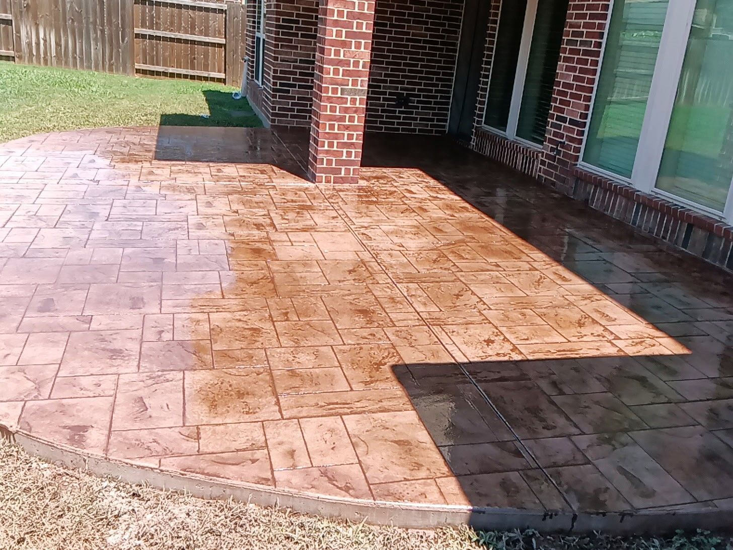 Stamped concrete patio beside a brick house, with a wooden fence and wet surface in sunlight.