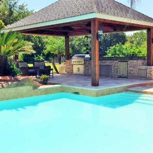 Poolside outdoor kitchen with a grill, under a wood-framed roof.