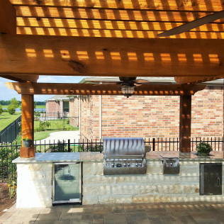 Outdoor kitchen with grill, refrigerator, and pergola. Brick building and lawn in background.