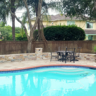 Swimming pool with a patio and table set, surrounded by a stone wall and wooden fence, a house in the background.