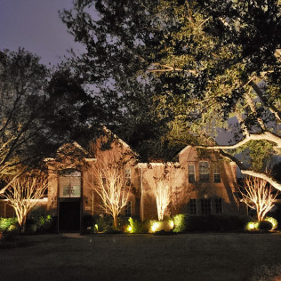 Illuminated house and trees at night with spotlights casting warm light.