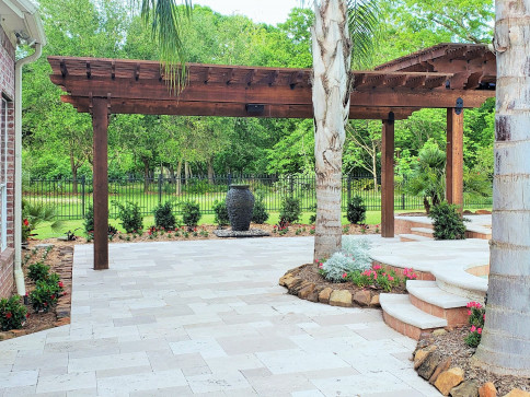 Patio with stone pavers, wooden pergola, and landscaping, featuring a fountain and steps.
