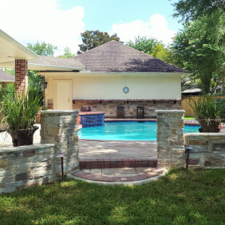 Backyard pool with stone walls and a building with a brown roof; potted plants and blue water.