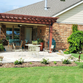 Patio with pergola, brick wall, white chairs and table, landscaping, and green lawn.