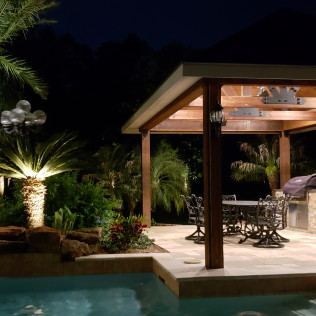 Nighttime view of a backyard with a pool, patio with pergola, and landscape lighting highlighting palm trees.