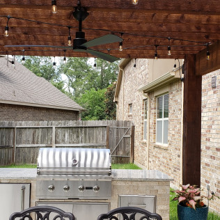 Outdoor kitchen with a grill, ceiling fan, and string lights under a wooden pergola.
