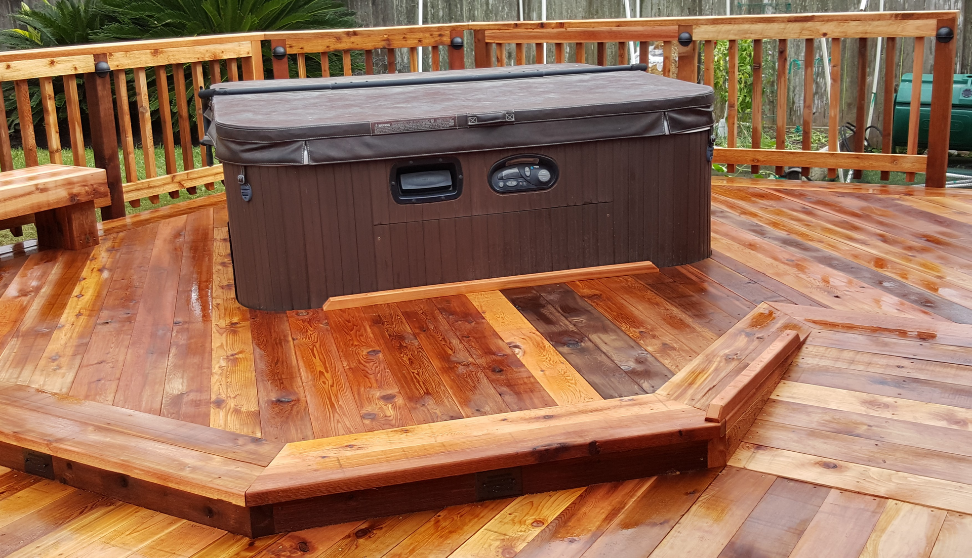 Hot tub with cover on a wet wooden deck, surrounded by a railing.