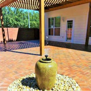 A water fountain on a brick patio under a wooden pergola, near a white building with a door.