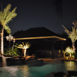 Nighttime view of a pool with waterfall, illuminated palm trees, and a gazebo.