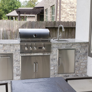 Outdoor kitchen with stainless steel grill, sink, and cabinets, against a stone facade.