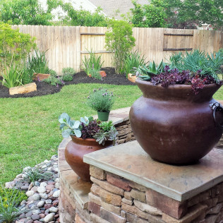 Two large brown pots with succulents on a stone patio in a backyard with a wooden fence.
