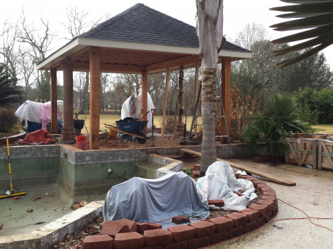 Gazebo construction near a drained pool; workers, building materials, and cloudy sky.