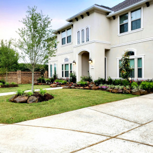Two-story beige house with manicured lawn and garden beds, concrete driveway.