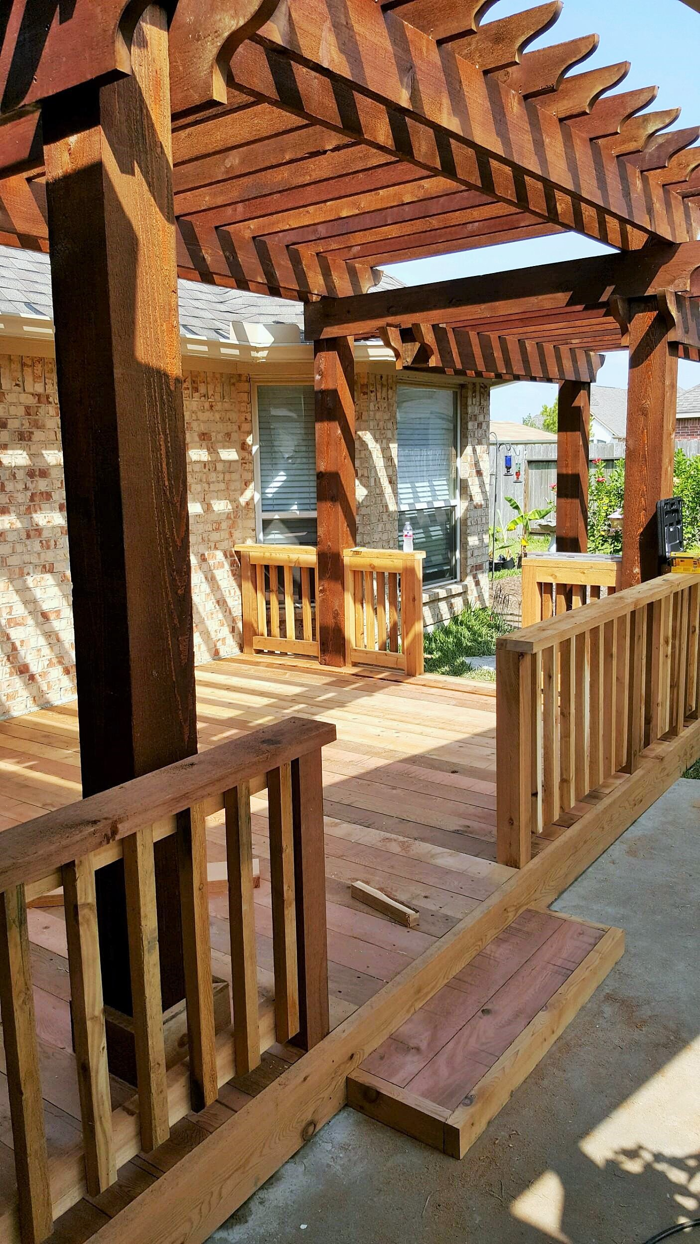 Wooden pergola over a deck with railings. Brown wood, bright daylight, small structure near a house.