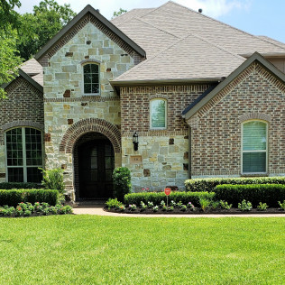 House exterior with stone and brick facade, arched doorway, green lawn, manicured hedges.