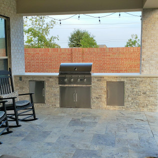 Outdoor kitchen with built-in grill, doors, and stone facade; brick wall in the background; string lights overhead.