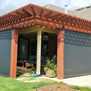 Outdoor patio with dark gray shades and a wooden pergola, lush greenery, and brick.