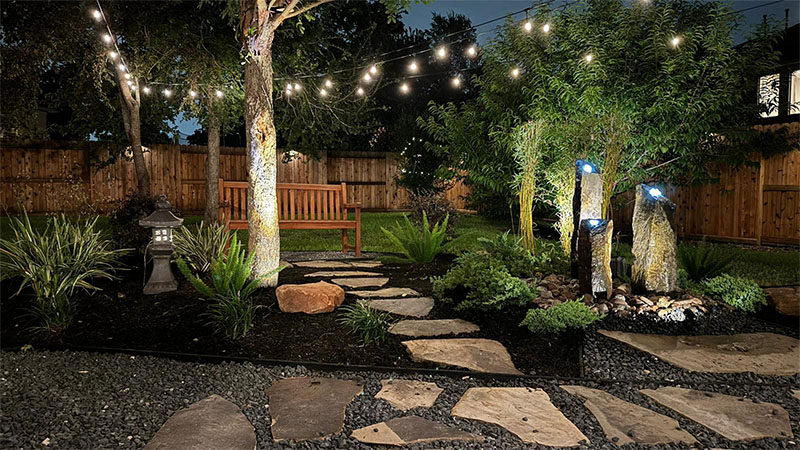 Backyard at night, stone path, string lights, bench, garden features, trees, illuminated rocks, and dark gravel.