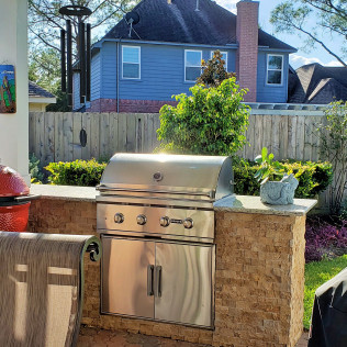 Stainless steel outdoor grill on a stone counter, against a fence and blue house.