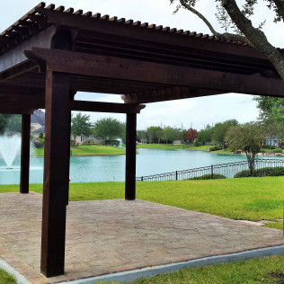Pergola overlooking a lake with fountain. Dark wood structure, concrete patio, green lawn, and trees.