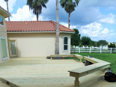Wooden deck with built-in bench, connecting to a house with red tile roof, and a white fence in the background.