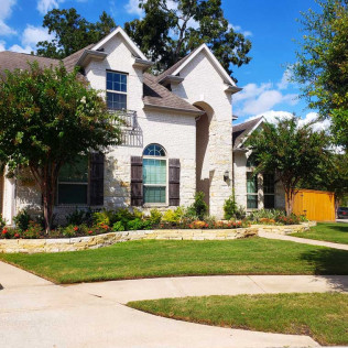 Two-story white brick house with brown roof, green lawn, trees, and blue sky.