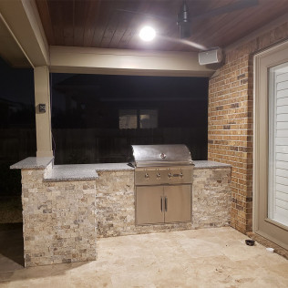 Outdoor kitchen with stainless steel grill, stone facade, and overhead lighting.