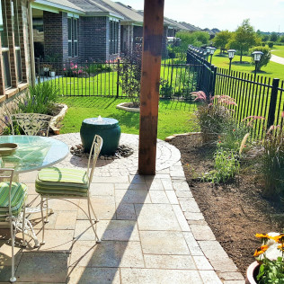 Patio with stone pavers, table, fountain, lawn, and black fence.