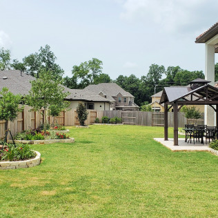 Lush green backyard with lawn, flowerbeds, wooden fence, gazebo with table and chairs, and surrounding houses.