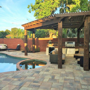 Patio with pool, pergola, outdoor kitchen, and seating area. Brick wall backdrop, paved ground.