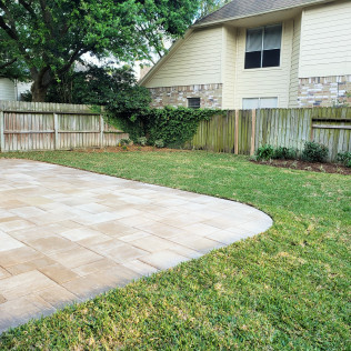 Stone patio with curved edge, bordered by grass, wooden fence, and a two-story beige house.