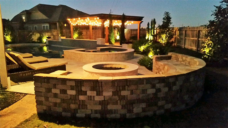 Backyard patio with fire pit, seating, and pergola with string lights, at dusk.