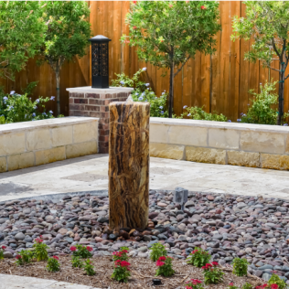 Water fountain with a textured column, surrounded by rocks and flowers, in a landscaped garden.