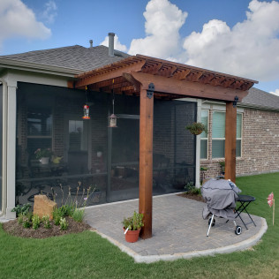 Patio with pergola, screen enclosure, grill, and landscaping next to a brick house on a sunny day.