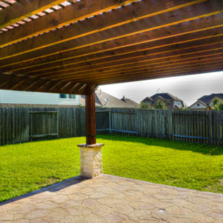 Backyard patio with wooden pergola, stone flooring, green grass, and wooden fence.
