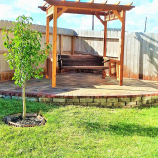 Wooden pergola with swing seat on a brick patio, next to a tree, in a backyard.