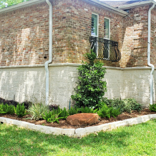 Corner of a brick house with a garden bed featuring plants and a large rock, on a green lawn.