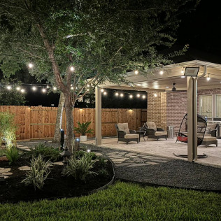 Backyard with string lights, pergola, seating area, tree, and wooden fence at night.